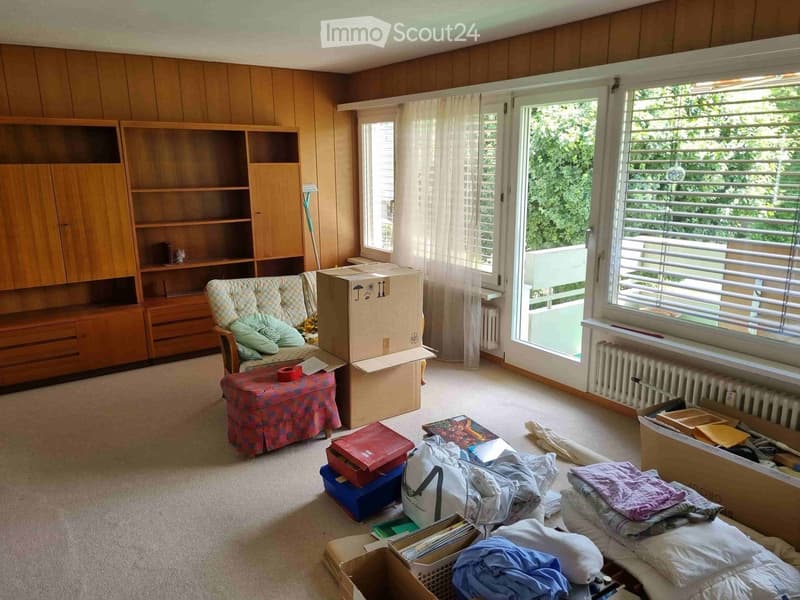 Living room with wooden panel walls, large windows, a sofa with cushions, a bookcase, a radiator, and several boxes and items scattered around.