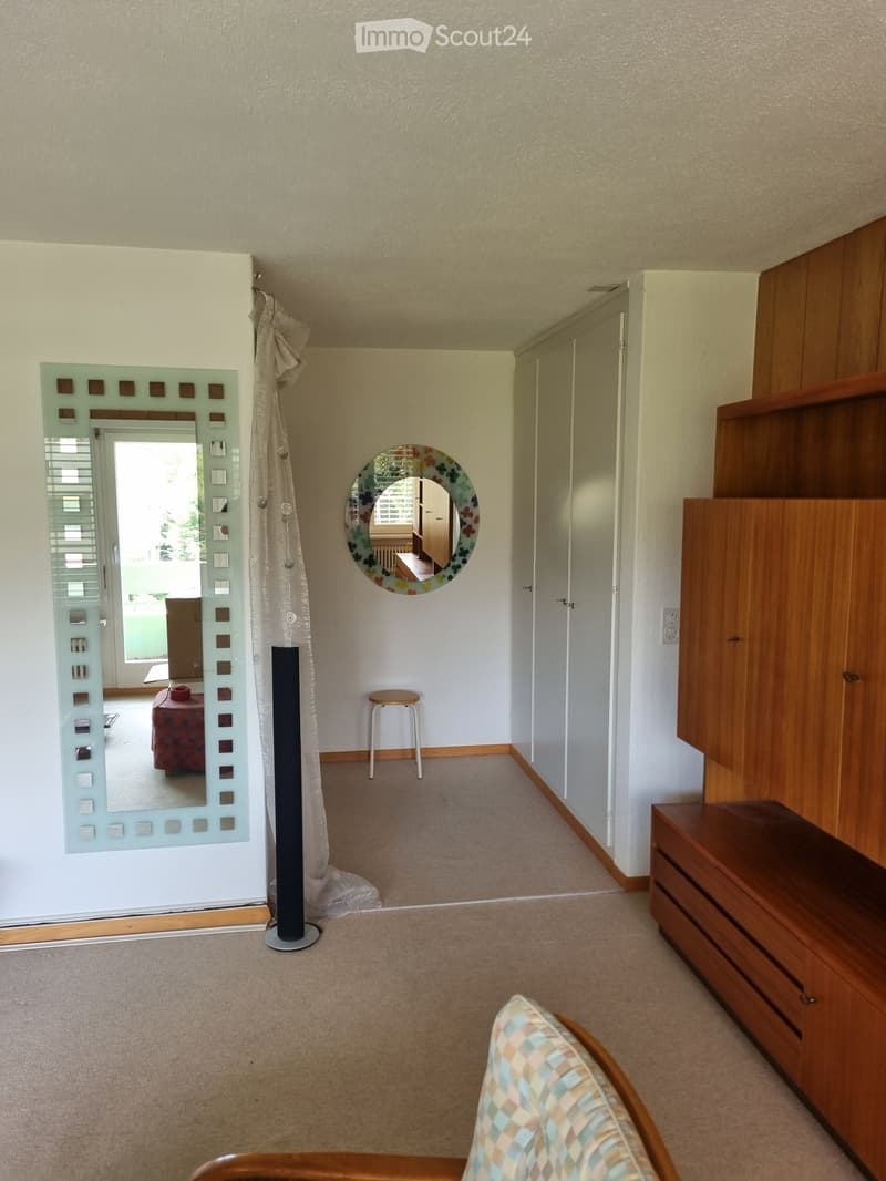 A small, well-lit room with a wooden cabinet, a patterned chair, a round mirror, a floor lamp, and a curtain. A small stool is near the doorway.
