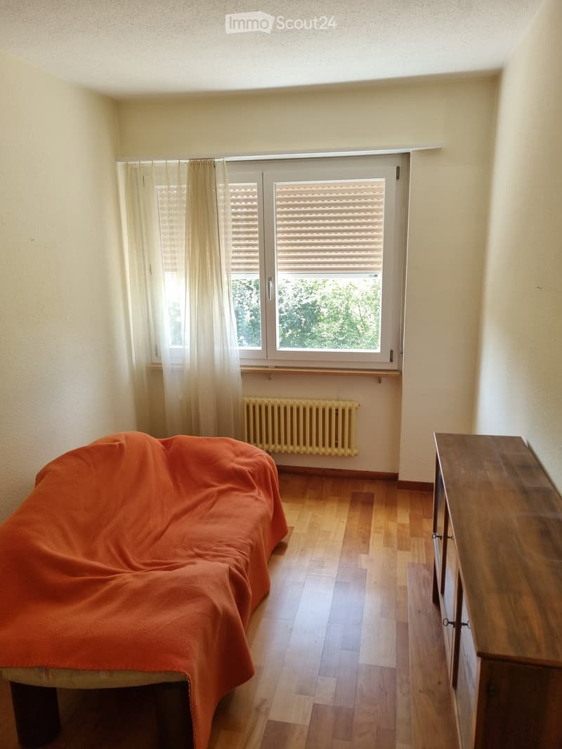 Bedroom with wooden flooring, a bed covered in an orange blanket, a radiator, a dresser, and a window with blinds and curtains.