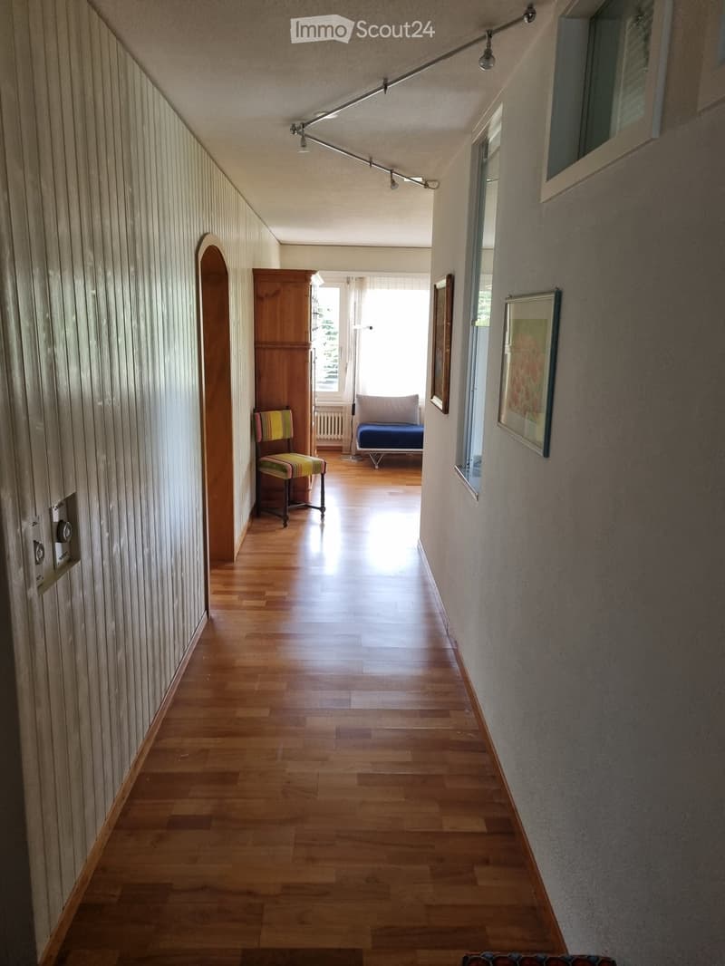 Wooden floor hallway with light-colored walls, a track light fixture, and a small seating area visible at the end.