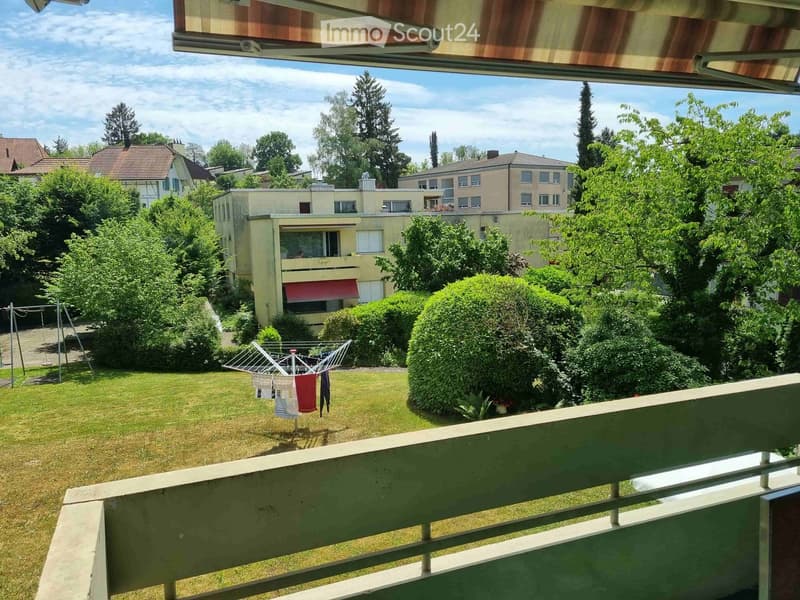 Balcony overlooking a green garden area with a clothesline, trees, and neighboring buildings.