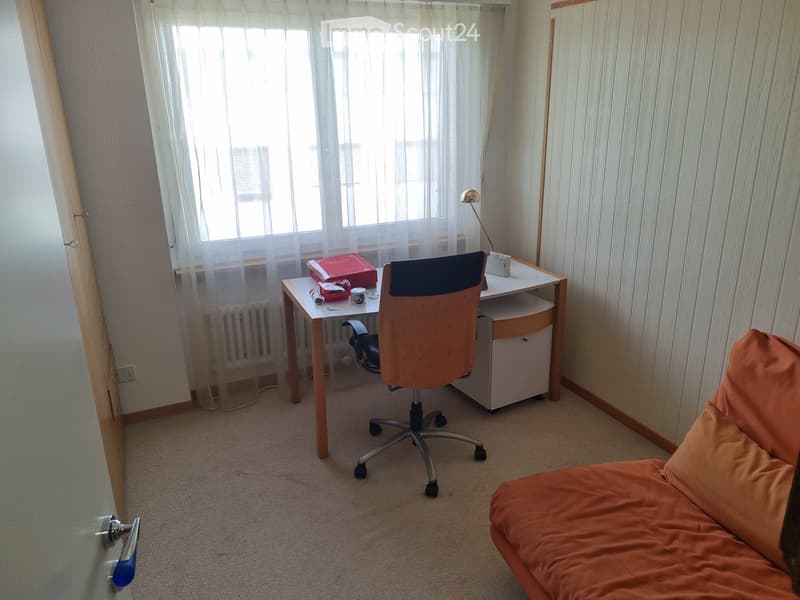 A small, well-lit room with a sofa, a desk, a chair, and a lamp. The room has a window with sheer curtains and a radiator below it.