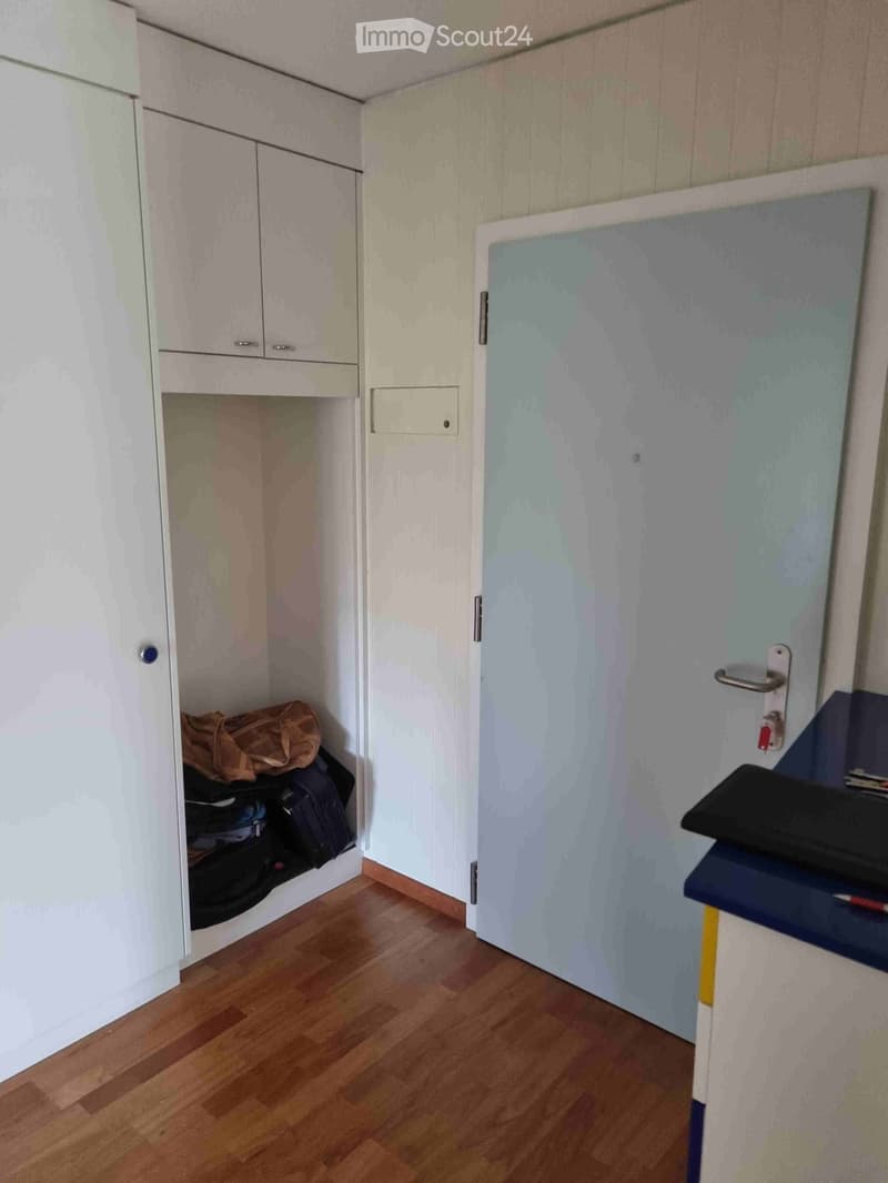 A small, furnished room with a wooden floor, white built-in closet, and a desk with a laptop on it. The closet contains a suitcase and other items.