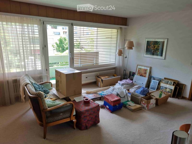 Living room with a sofa, small table, chair, picture frames, and a floor lamp. The room is partially packed with boxes and items, indicating a move or storage.