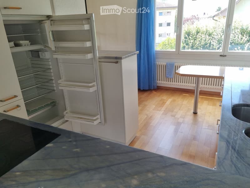 Empty kitchen with wooden flooring, a refrigerator, a small table, and a sink visible.