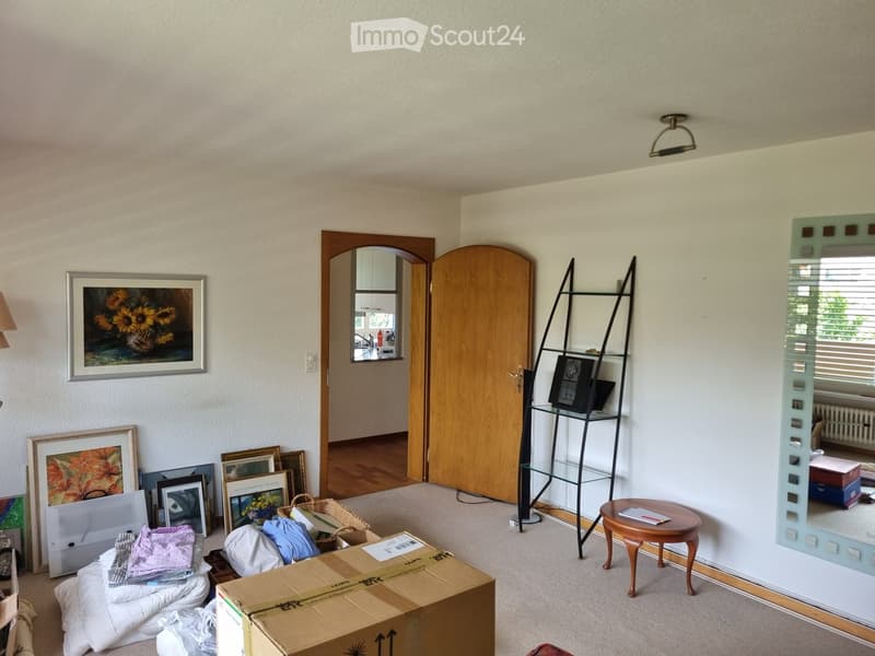Living room with a round wooden table, a tall black shelving unit, a large mirror, several picture frames, and a basket on the floor.