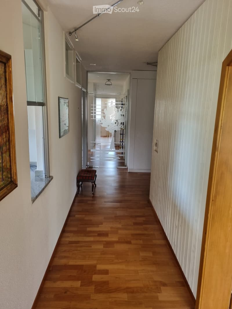 A long, narrow hallway with wooden flooring, white walls, and recessed lighting. A small bench sits against the left wall, and a glass door with a decorative pattern leads to another room.