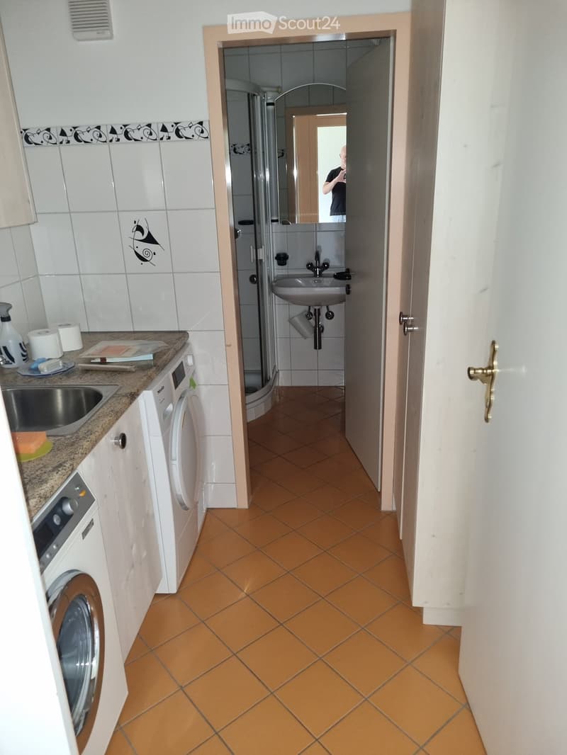 Hallway with orange tiled floor leading to a bathroom and a kitchen area. The bathroom has a sink, mirror, and shower stall, while the kitchen features a washing machine, sink, and countertop with various items.