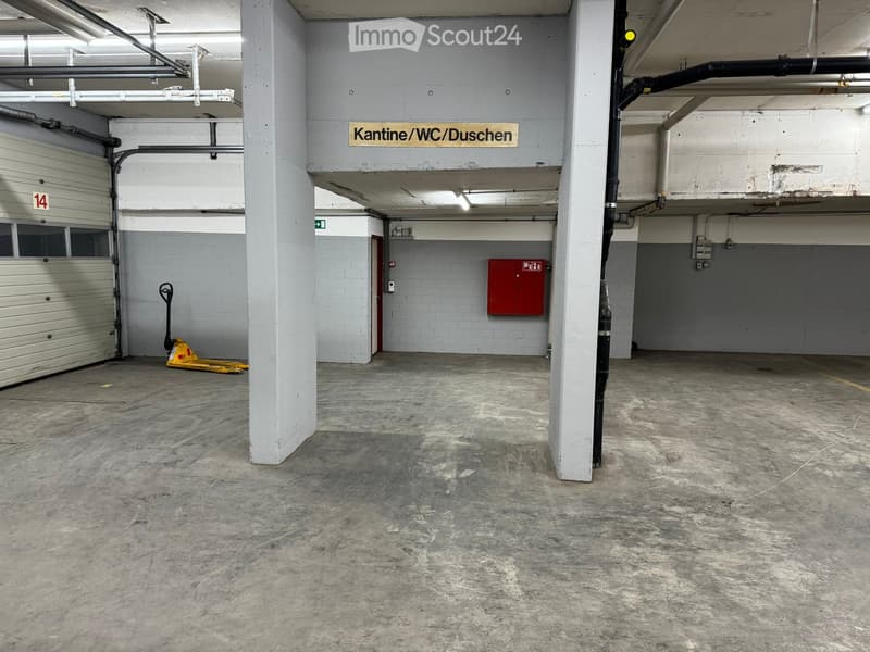 An empty underground parking garage with white concrete walls and a ceiling, a door to the right, a red storage cabinet next to the door, and a sign on the wall that reads 'Kantine/WC/Duschen'.