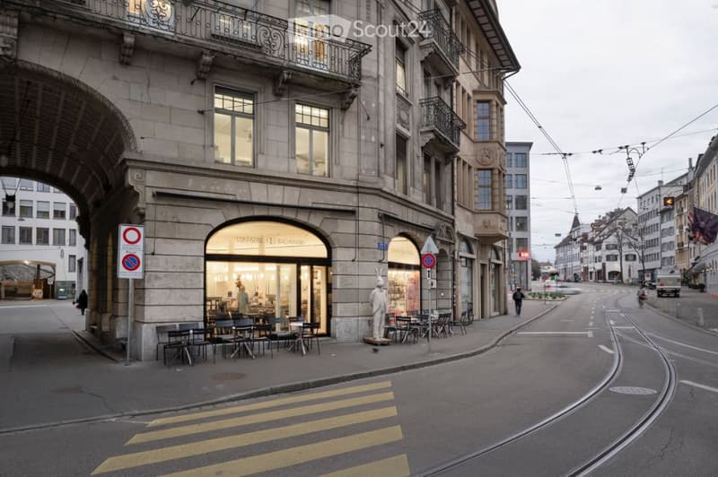 Urban street view with a multi-story building featuring large windows, balconies, and a storefront with outdoor seating. The street includes traffic signs, a pedestrian crossing, and a tram line.