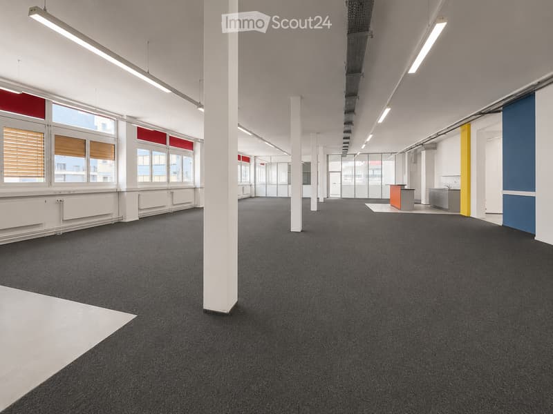 Open-plan office space with dark carpeted flooring, white walls, large windows with blinds, and minimal furniture including a small reception desk and a sink area.