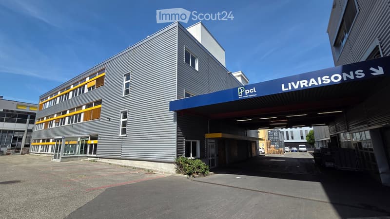 Modern industrial-style building with grey metal facade, large windows, and a blue sign reading "LIVRAISONS" above the entrance.