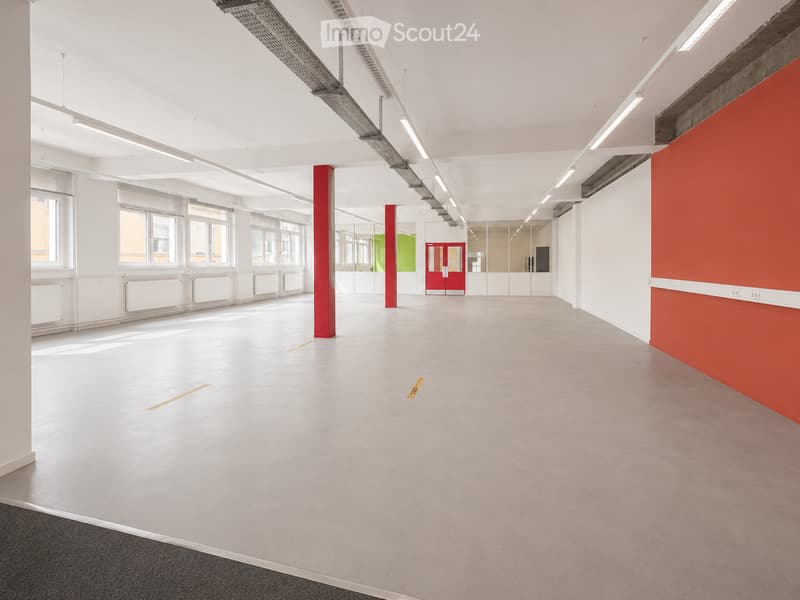 Empty room with white walls, grey floor, red pillars, and large windows allowing natural light. No furniture or equipment present.