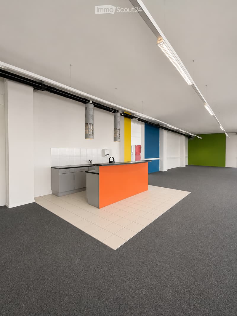 Modern, open-plan room with a grey carpeted floor, white tiled backsplash, grey cabinets, and an orange counter. The space features colorful accent walls in yellow, blue, and green, with pendant lighting fixtures.