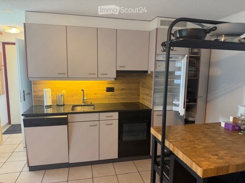 Modern kitchen with light-colored cabinets, a black countertop, and built-in appliances including a dishwasher, oven, and refrigerator. A wooden island with a pot and bottle is visible.