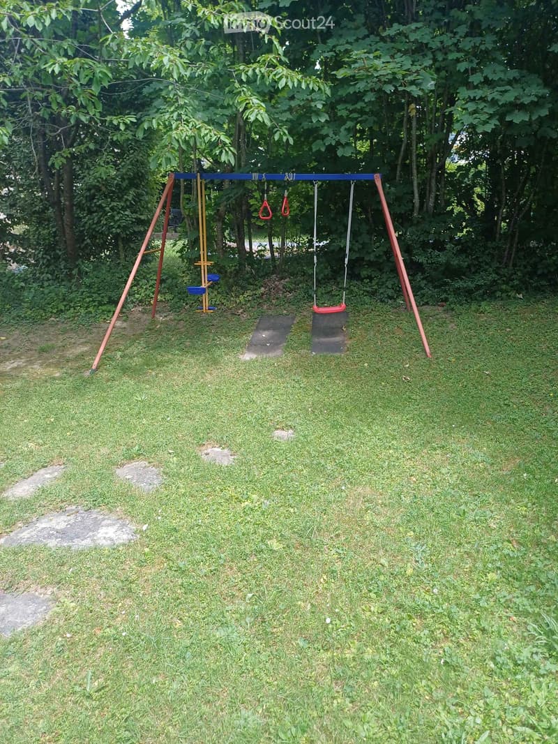 A grassy backyard with a swing set featuring swings and monkey bars, surrounded by trees and shrubs.