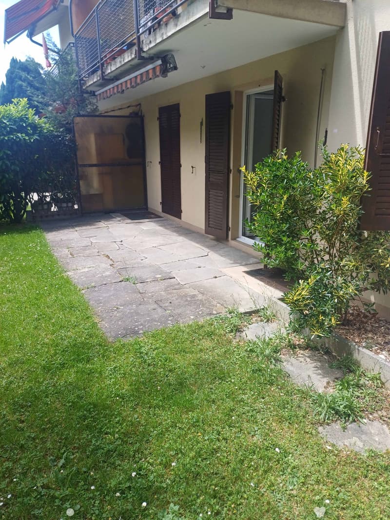 Garden area with a stone pathway leading to a house entrance, featuring a small grassy lawn, a few bushes, and a balcony with railings visible above.