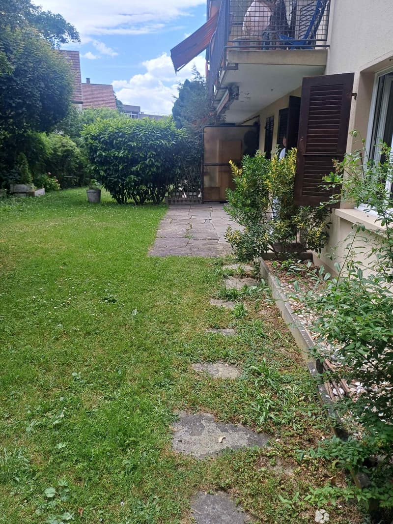 Garden area with a grassy lawn, concrete pathway, and various plants and shrubs. A building with a balcony is visible in the background.