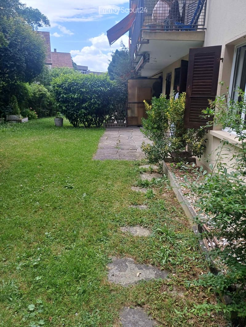 Garden area with a grassy lawn, concrete pathway, and various plants and shrubs. A building with a balcony is visible in the background.