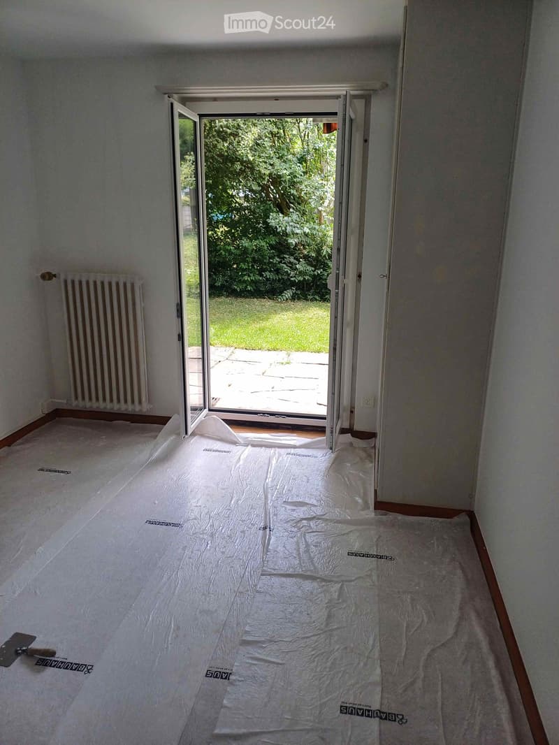 Empty room with white plastic sheeting on the floor, a radiator on the left wall, and an open sliding door leading to a green outdoor area.