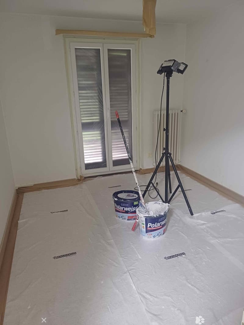 Empty room with bare walls and a plastic drop cloth on the floor, featuring a tripod with lights, paint buckets, and a paint roller.