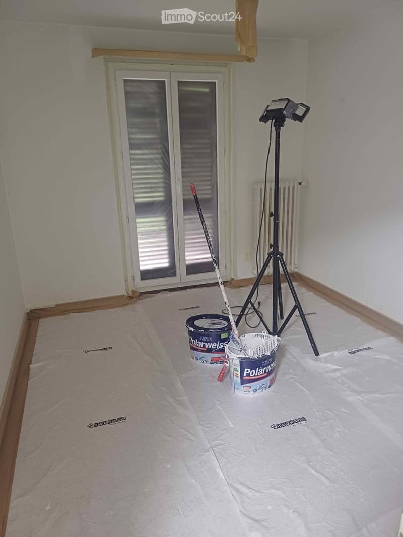 Empty room with bare walls and a plastic drop cloth on the floor, featuring a tripod with lights, paint buckets, and a paint roller.