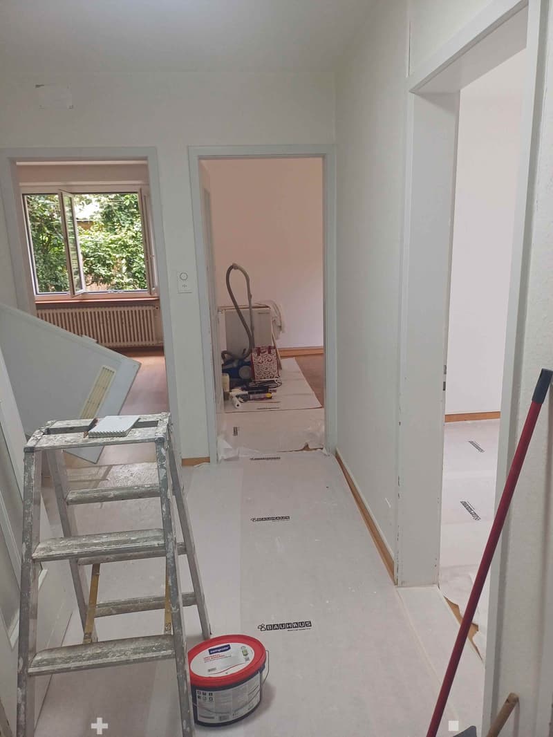 Empty hallway with white walls, a ladder, a paint can, and a broom. A partially open door leads to another room with a bed and cleaning supplies.
