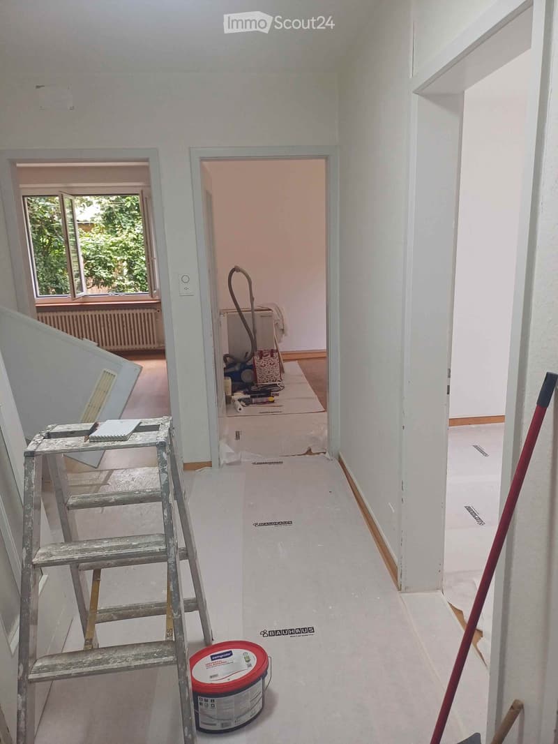 Empty hallway with white walls, a ladder, a paint can, and a broom. A partially open door leads to another room with a bed and cleaning supplies.