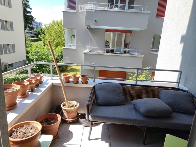 Balcony with a grey wicker sofa, several terracotta pots, and a broom leaning against the railing.