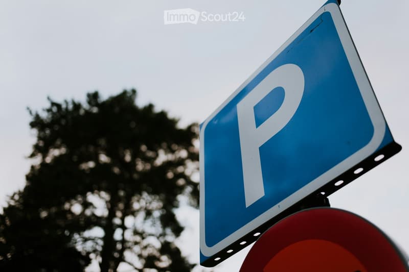 A blue parking sign with a white 'P' symbol, mounted on a pole, with a blurred background of trees and a red circular sign below.