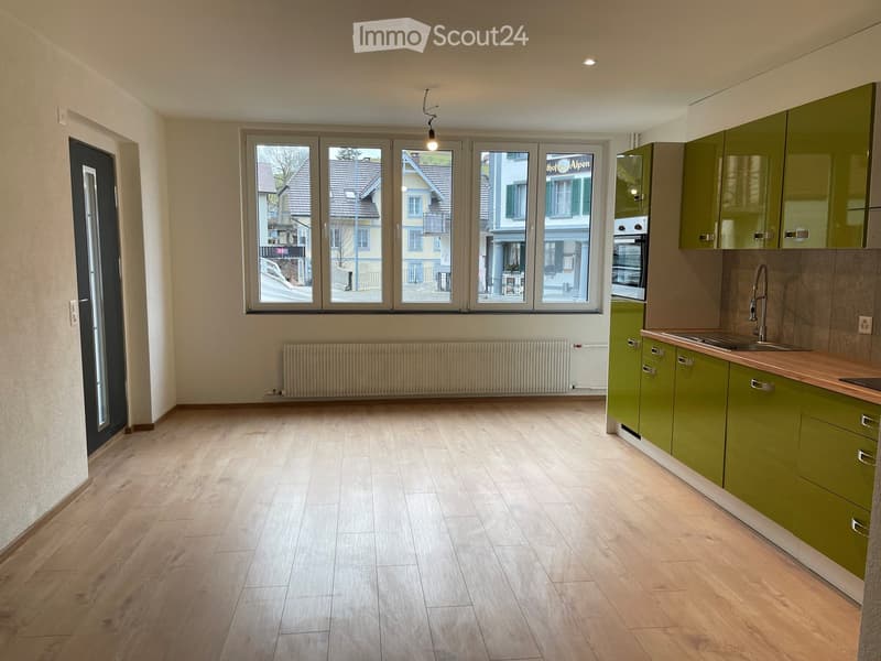 Empty kitchen with light wooden flooring, green cabinets, a sink, and large windows overlooking a street.