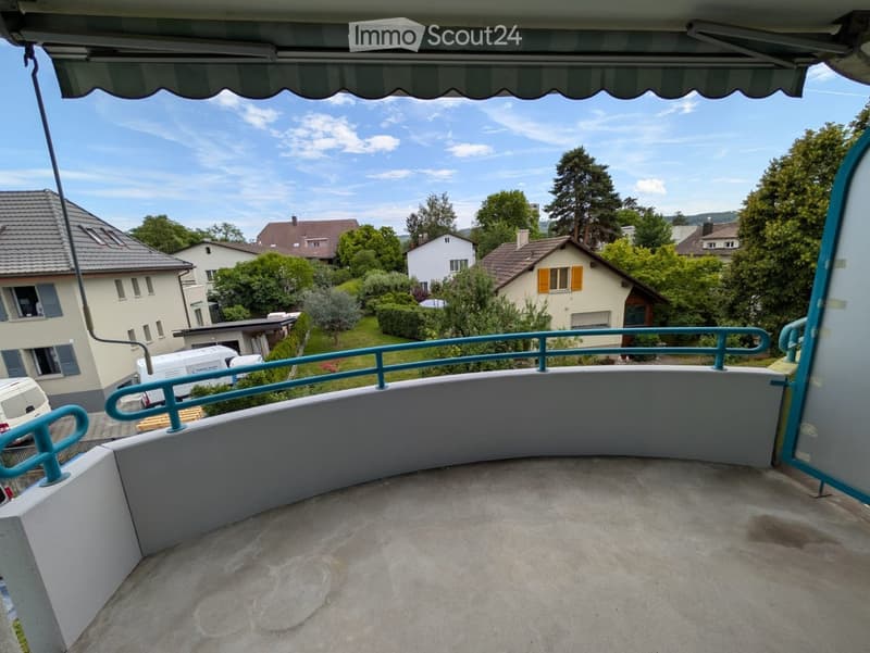 Balcony with a concrete floor, turquoise railing, and a striped awning, overlooking a residential area with houses and greenery.