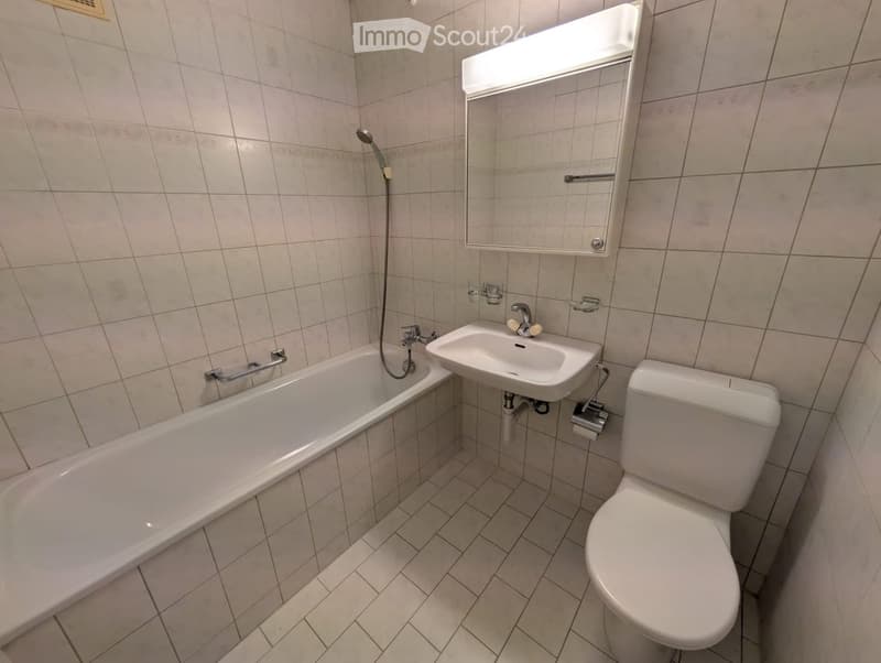 Bathroom with a bathtub, sink, toilet, mirror, and showerhead. The walls and floor are tiled in a light beige color.