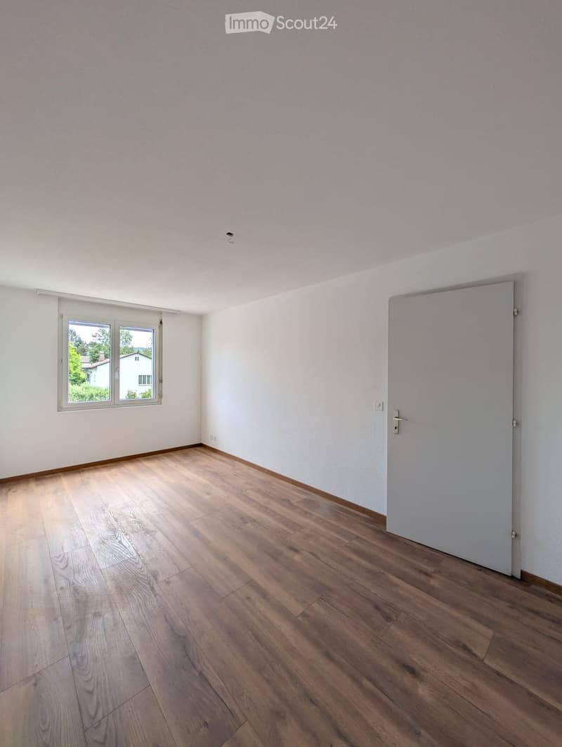 Empty room with light wooden flooring, white walls, and a large window with a view of the outside. There is a closed door on the right side.
