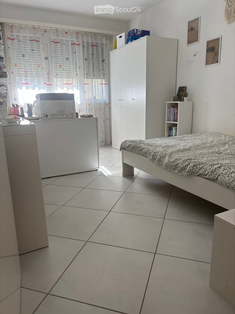 A tidy bedroom with a bed, a wardrobe, a desk with a printer, a bookshelf, and a window with curtains.