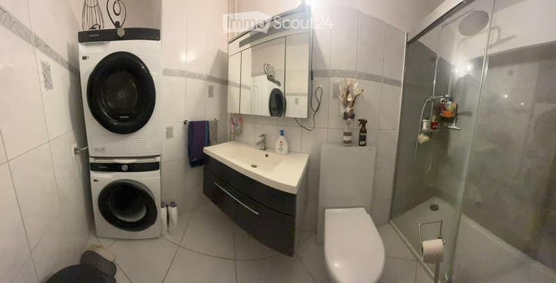 Modern bathroom with a sink, mirror, toilet, shower, and a stacked washing machine. Includes a towel, decorative vase, and cleaning supplies.