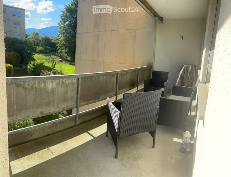 Balcony with wicker chairs and a small table, overlooking a green park and distant mountains.