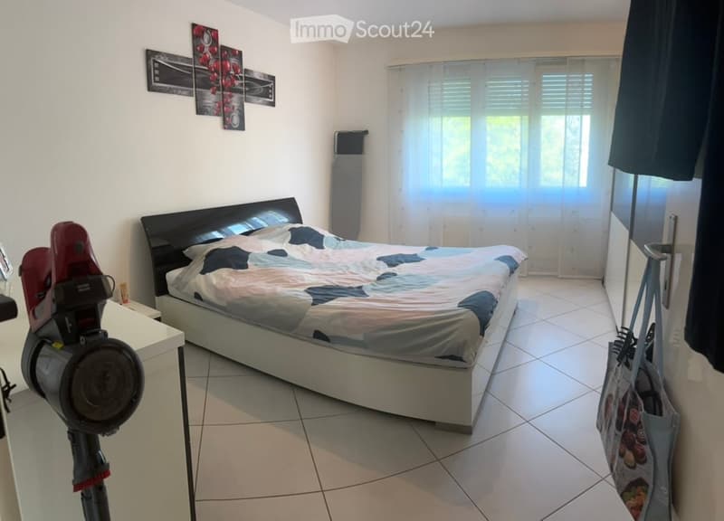Bedroom with a double bed, nightstand, vacuum cleaner, and handbag visible. The room has white tiled flooring and white walls with artwork.