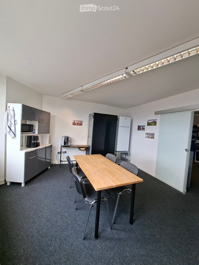 A modern, furnished room with a wooden table and chairs, a small kitchen area with a microwave, refrigerator, and coffee maker, and a desk with a computer. The room has grey carpeting and white walls.