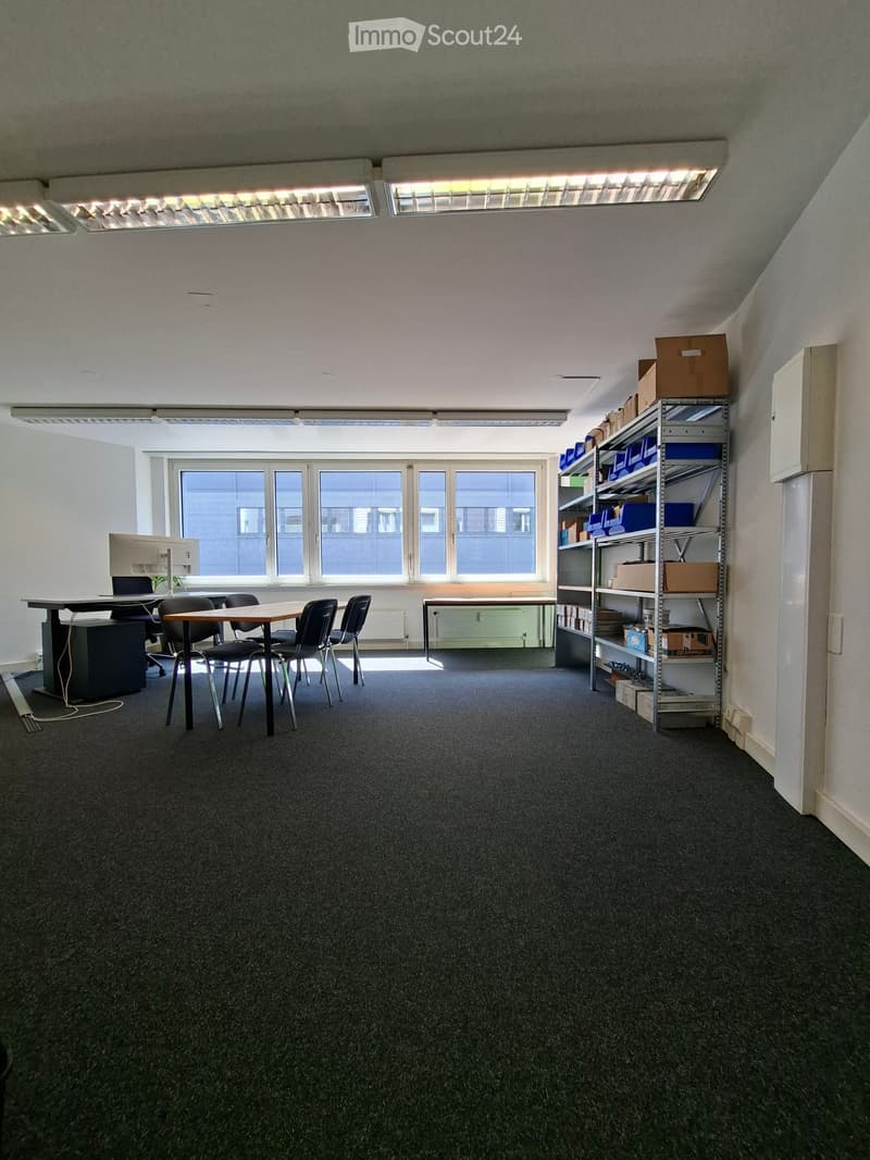 Office space with a table, chairs, a shelf with boxes, and a computer setup near a large window.