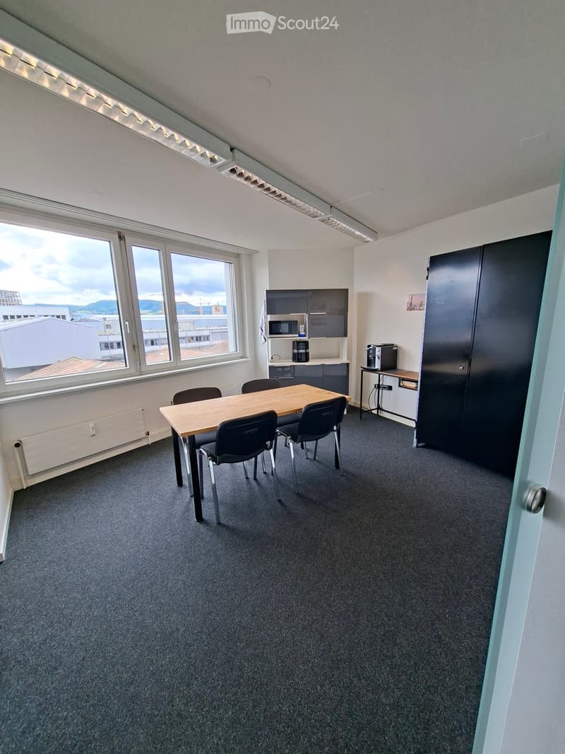 Bright room with large windows, a wooden table surrounded by chairs, a cabinet with a microwave, and a printer on a small table.