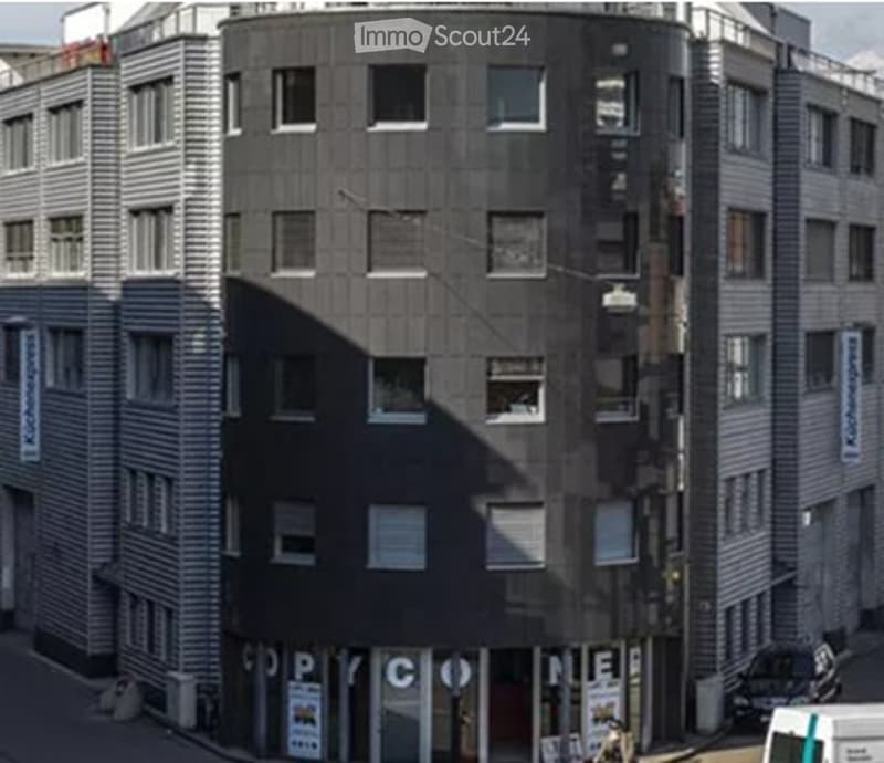 Modern multi-story building with a mix of grey and white facade, featuring numerous windows and a ground-floor commercial area with signage.