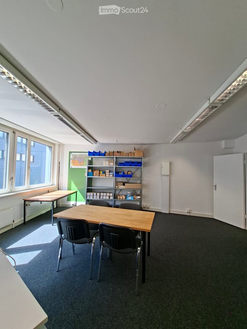 Bright room with wooden tables, black chairs, and a metal shelving unit holding boxes and storage bins. Large windows provide natural light.