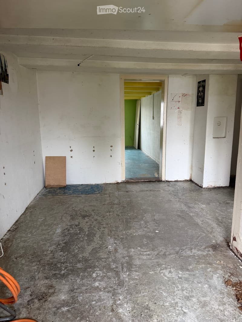 Empty hallway with concrete floor, white walls, and a doorway leading to another room with green walls and blue flooring.
