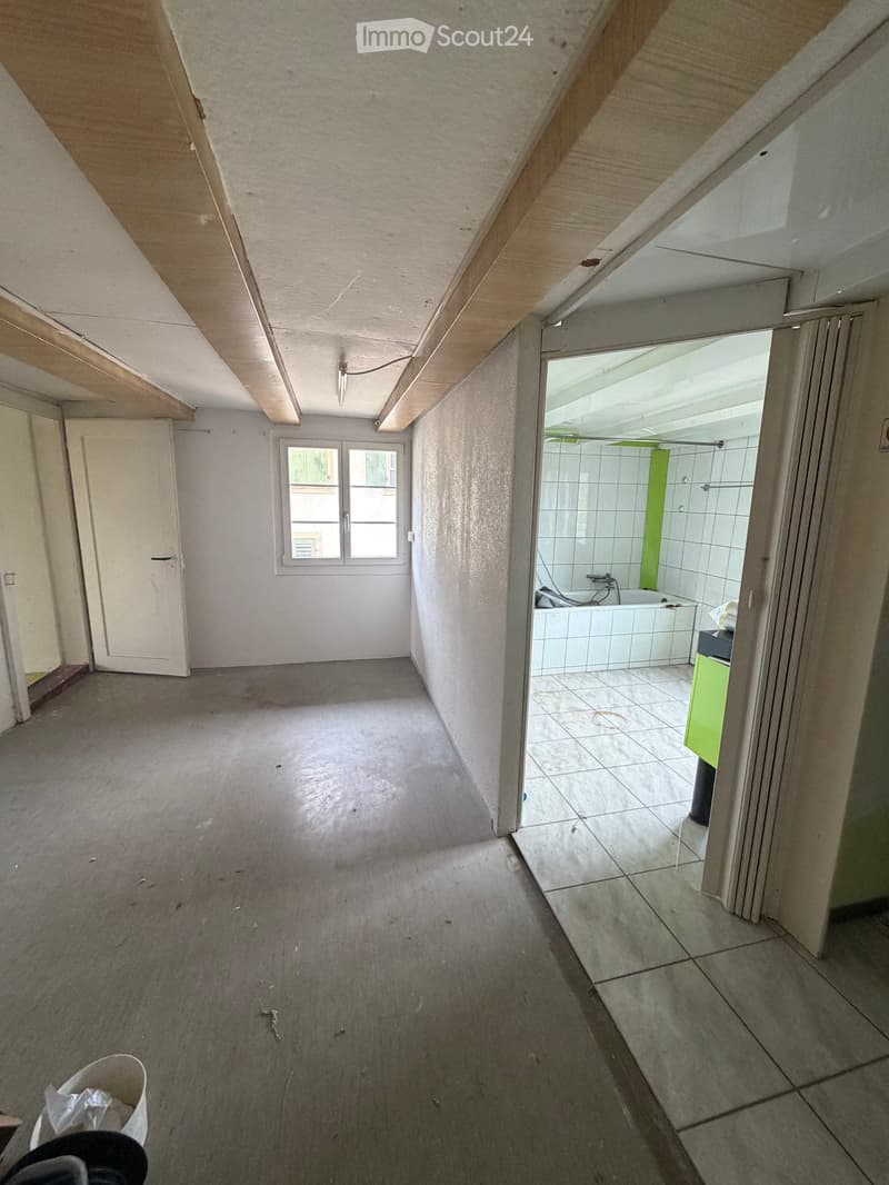 Empty room with concrete floor, white walls, and a window. Adjacent bathroom with tiled walls and a bathtub is visible.