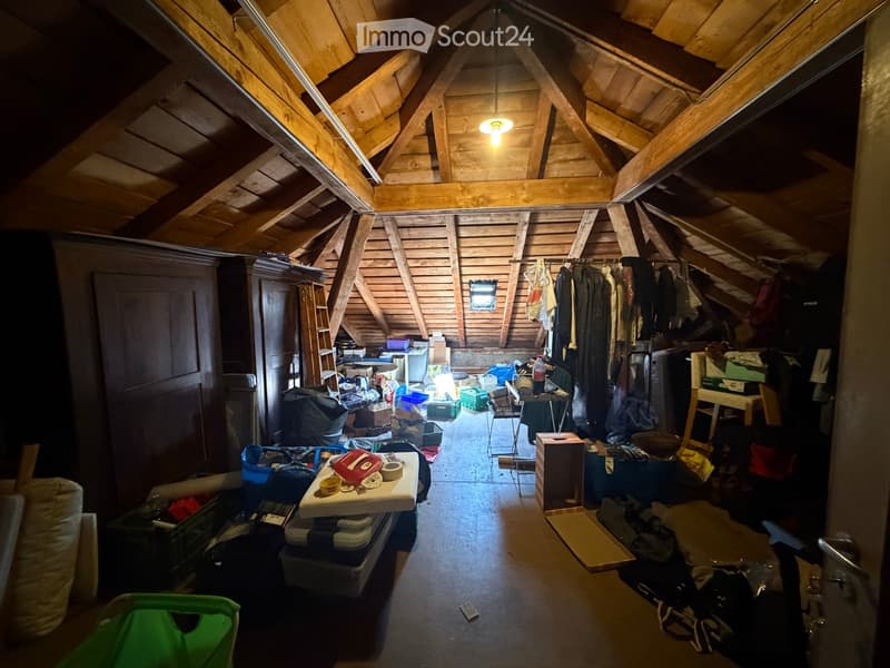 Attic space with wooden beams and exposed rafters, containing a small table with items, a clothing rack, storage boxes, and a ladder leading to another level.