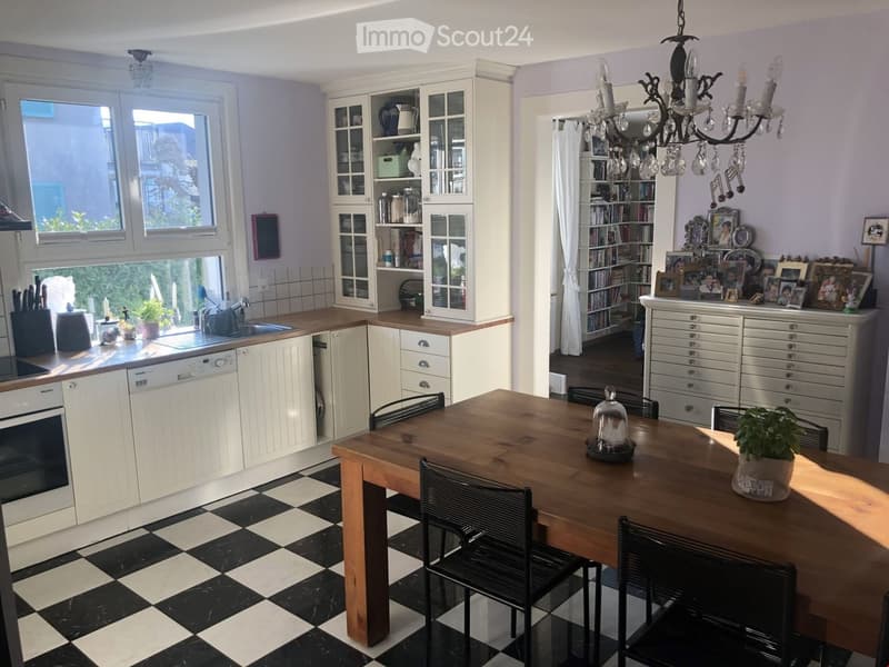 A well-lit kitchen and dining area with white cabinets, a wooden dining table, black chairs, a chandelier, and a bookshelf filled with books and decorative items.