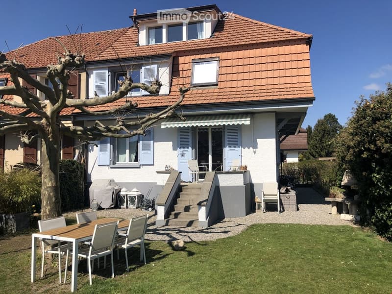 A two-story house with a red-tiled roof and white walls, featuring a well-maintained garden with a wooden table and chairs, steps leading to a covered patio area, and various outdoor furniture.