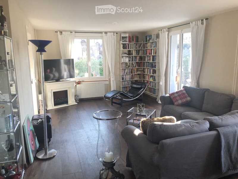 Modern living room with a grey sectional sofa, coffee table, glass side tables, a fireplace, a bookshelf, and a floor lamp. Large windows with white curtains provide natural light.