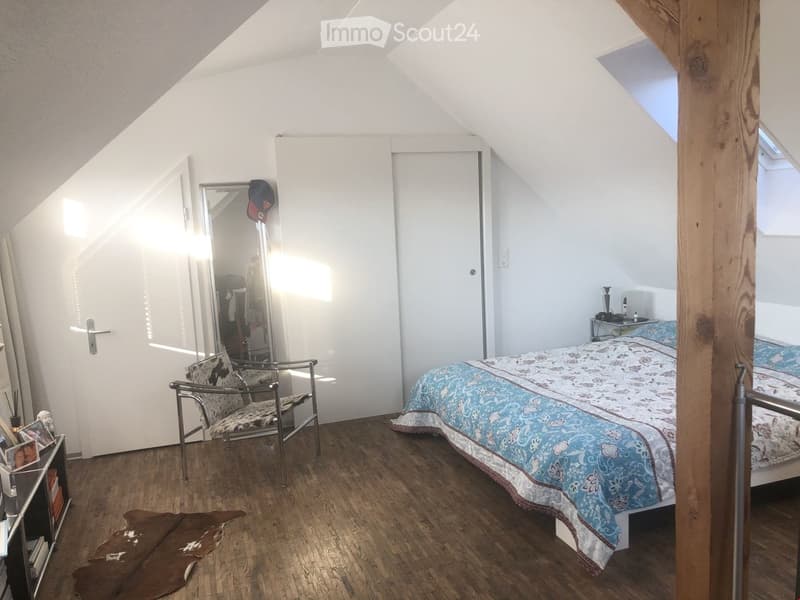 Cozy attic bedroom with wooden floors, slanted ceilings, and large windows. Features a double bed with blue patterned bedding, a metal-framed chair, a full-length mirror, and a small nightstand.